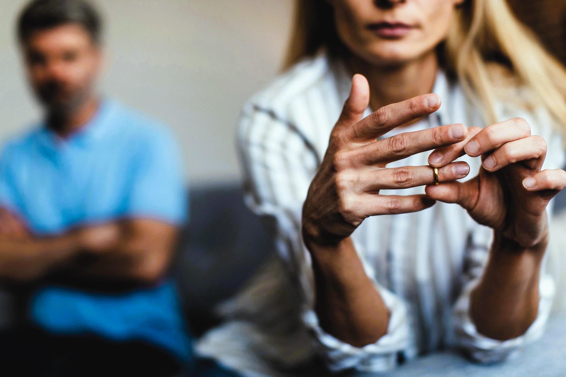 A woman sits in focus, looking down and removing a ring from her finger, symbolizing a separation or break-up. In the background, a man with crossed arms appears blurred, sitting on a couch. The scene conveys tension and emotional distance.