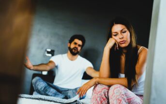 A woman sits on the edge of a bed looking contemplative and distant, while a man in the background gestures with his hands as if speaking or arguing. The room is dimly lit, creating a somber mood.