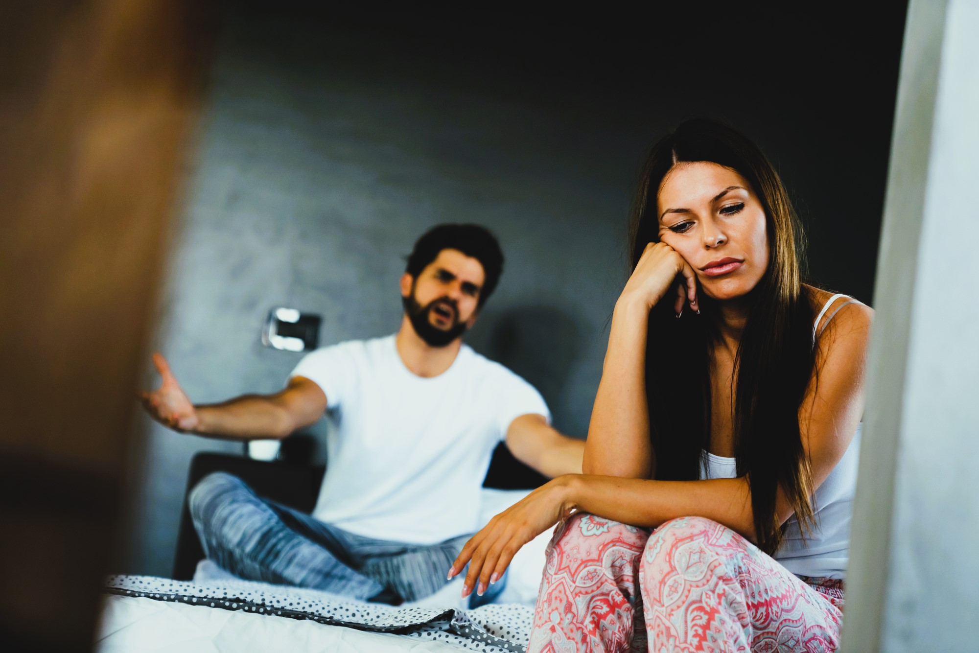 A woman sits on the edge of a bed looking contemplative and distant, while a man in the background gestures with his hands as if speaking or arguing. The room is dimly lit, creating a somber mood.