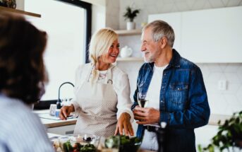 A woman and a man are in a kitchen, smiling and chatting. The woman is wearing an apron and pointing towards a bowl of vegetables, while the man holds a glass of wine. Another person is in the foreground, slightly blurred.