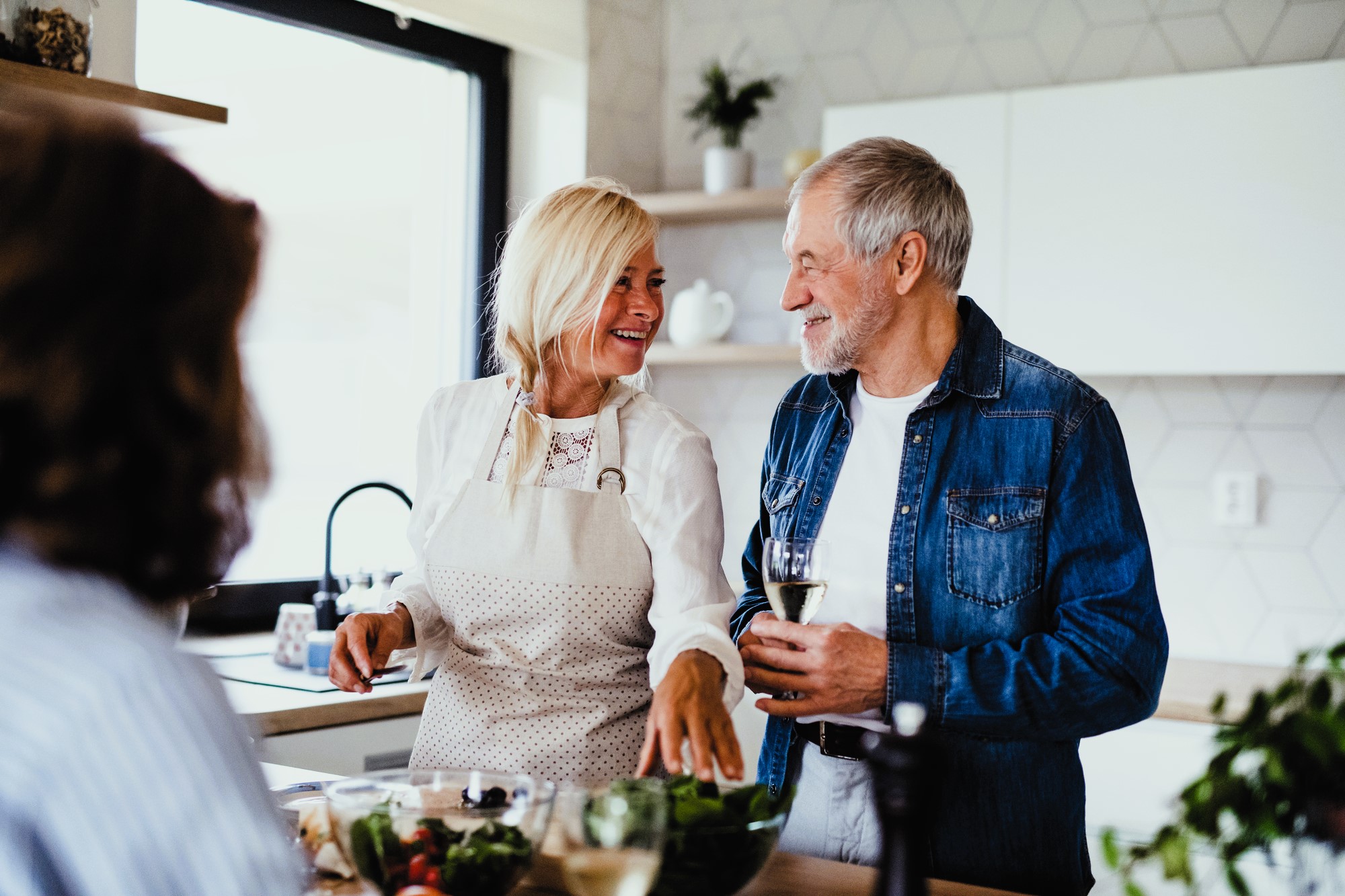 A woman and a man are in a kitchen, smiling and chatting. The woman is wearing an apron and pointing towards a bowl of vegetables, while the man holds a glass of wine. Another person is in the foreground, slightly blurred.