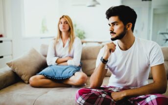 A man and woman sit on a beige couch in a brightly lit room. The man, wearing a white t-shirt and red plaid pants, appears thoughtful, resting his chin on his hand. The woman, in a white blouse and jeans, sits cross-legged with her arms folded, looking away.