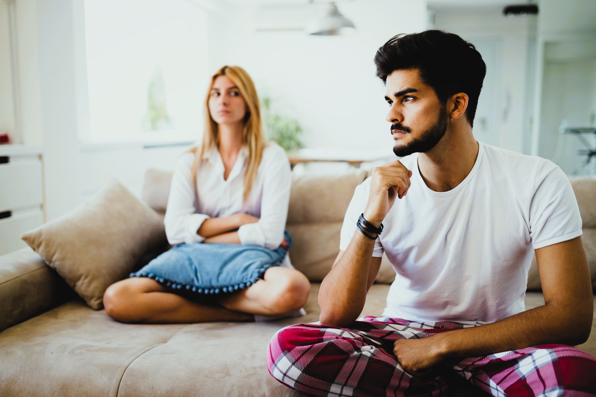 A man and woman sit on a beige couch in a brightly lit room. The man, wearing a white t-shirt and red plaid pants, appears thoughtful, resting his chin on his hand. The woman, in a white blouse and jeans, sits cross-legged with her arms folded, looking away.