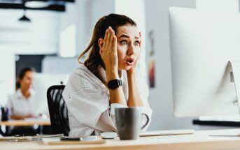 A woman with a ponytail looks shocked as she sits at a desk in front of a computer, her hands on her face. She is wearing a white shirt and a smartwatch. A coffee mug is on the desk. An office setting is visible in the background.