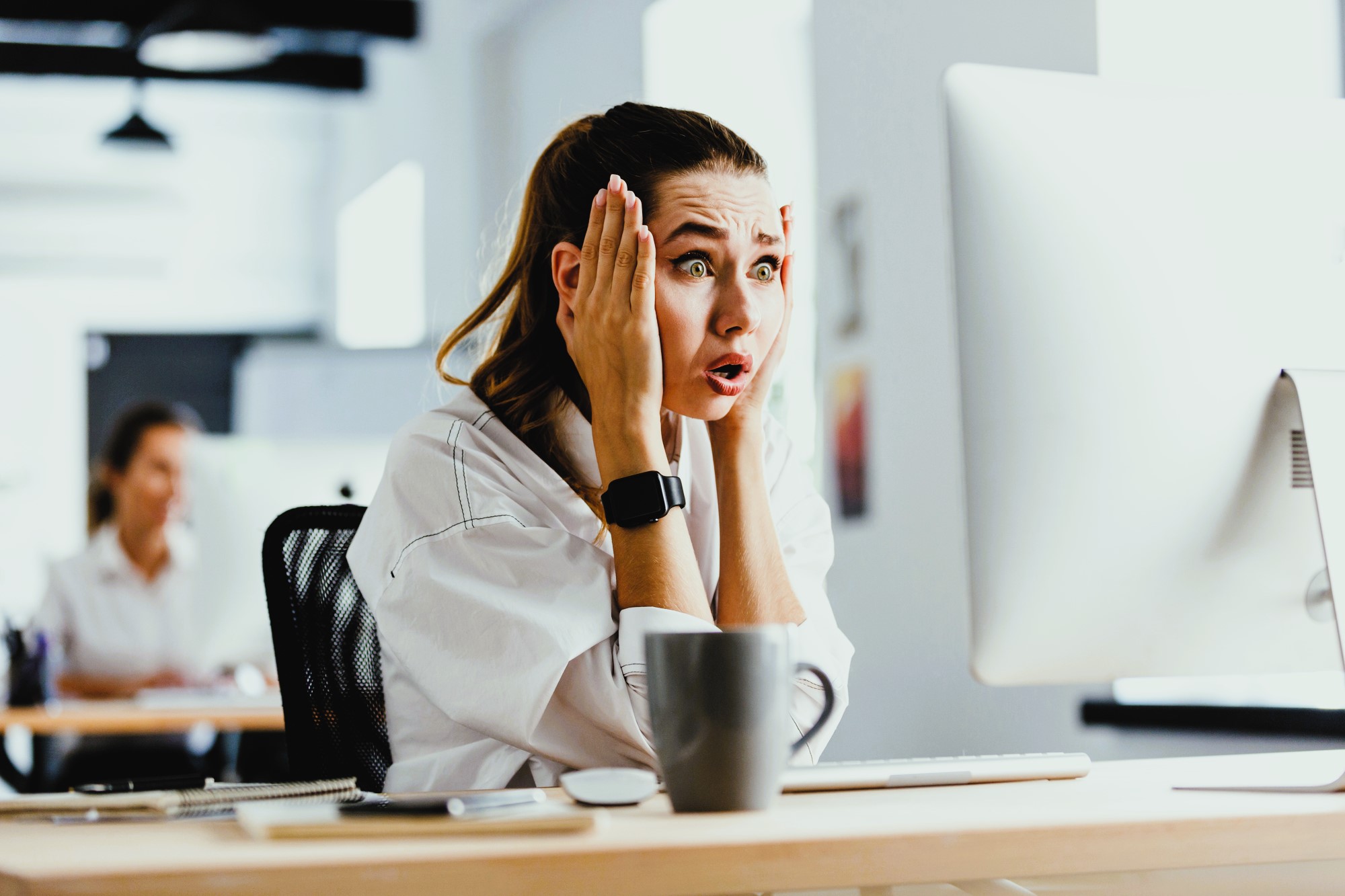 A woman with a ponytail looks shocked as she sits at a desk in front of a computer, her hands on her face. She is wearing a white shirt and a smartwatch. A coffee mug is on the desk. An office setting is visible in the background.