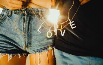 Two people stand close together, each holding a string with colorful letters spelling "LOVE" between them. They are wearing denim shorts and a black shirt, with sunlight shining through the letters. The mood is warm and intimate.