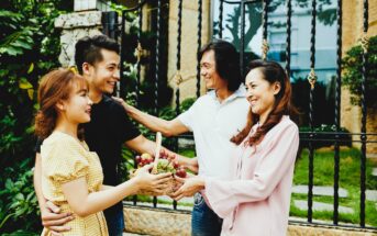 A young couple hands a basket of fruit to an older couple outdoors. Both couples are smiling warmly at each other in front of a wrought iron fence with greenery around them.