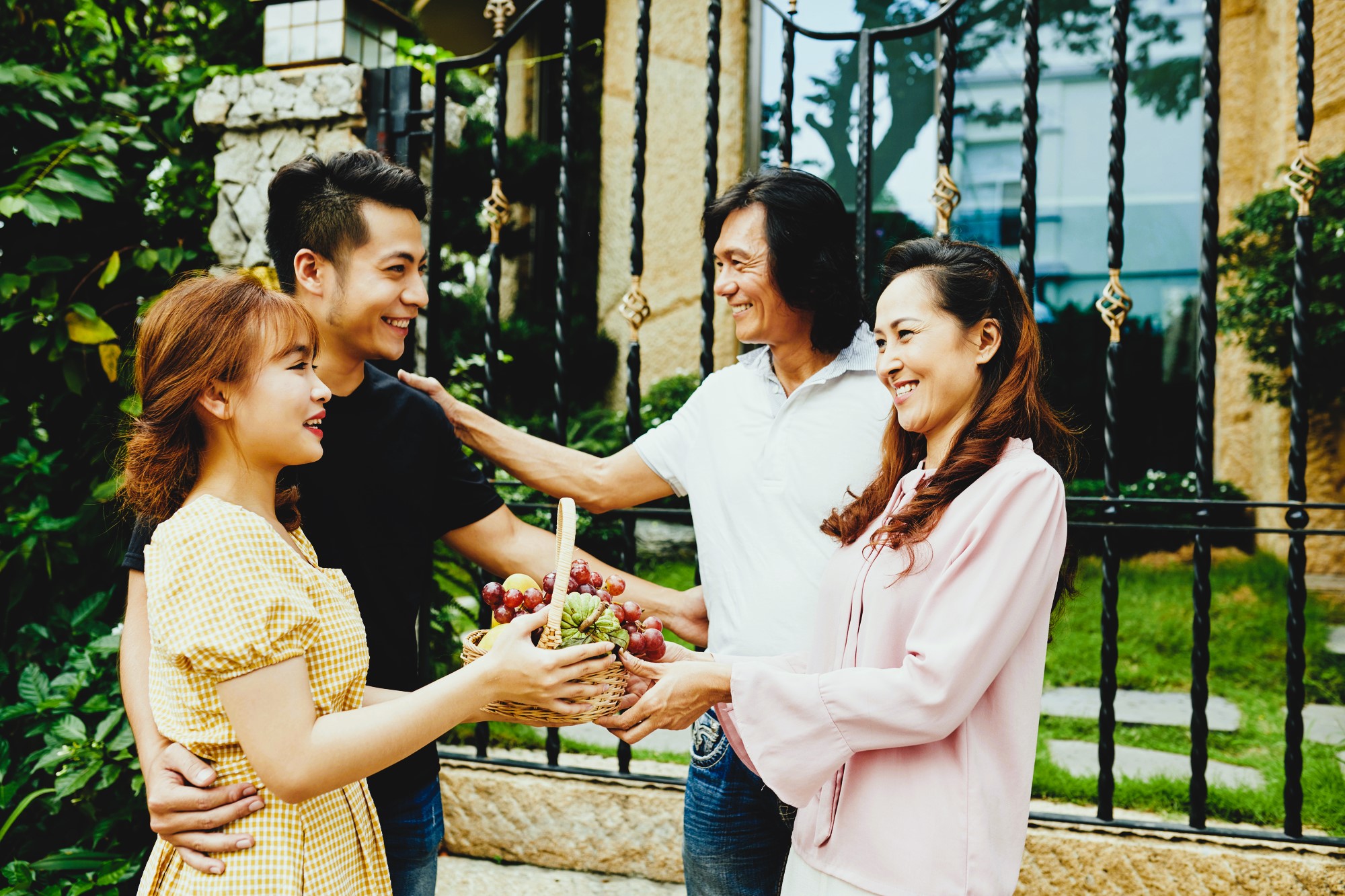 A young couple hands a basket of fruit to an older couple outdoors. Both couples are smiling warmly at each other in front of a wrought iron fence with greenery around them.