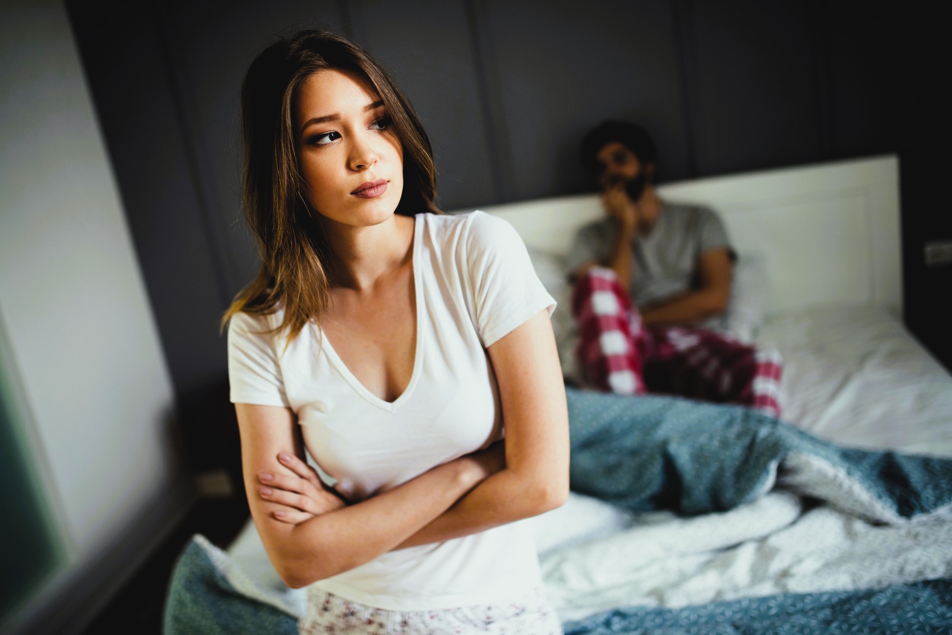 A woman with crossed arms stands at the forefront, looking away pensively. In the background, a man sits on a bed wearing pajamas, appearing thoughtful. The setting suggests a bedroom with a softly lit atmosphere.