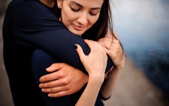 A woman is being gently embraced from behind by someone, their arms wrapped around her. She is wearing a black top and has her eyes closed, appearing peaceful. The background shows a blurred water body and pathway.