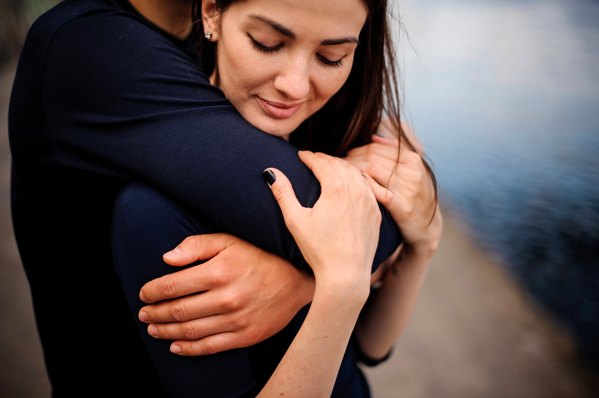 A woman is being gently embraced from behind by someone, their arms wrapped around her. She is wearing a black top and has her eyes closed, appearing peaceful. The background shows a blurred water body and pathway.