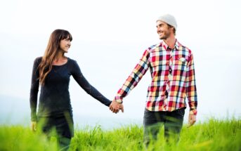 A woman in a dark dress and a man in a plaid shirt with a beanie hold hands, smiling at each other. They stand in a grassy field under a bright sky.