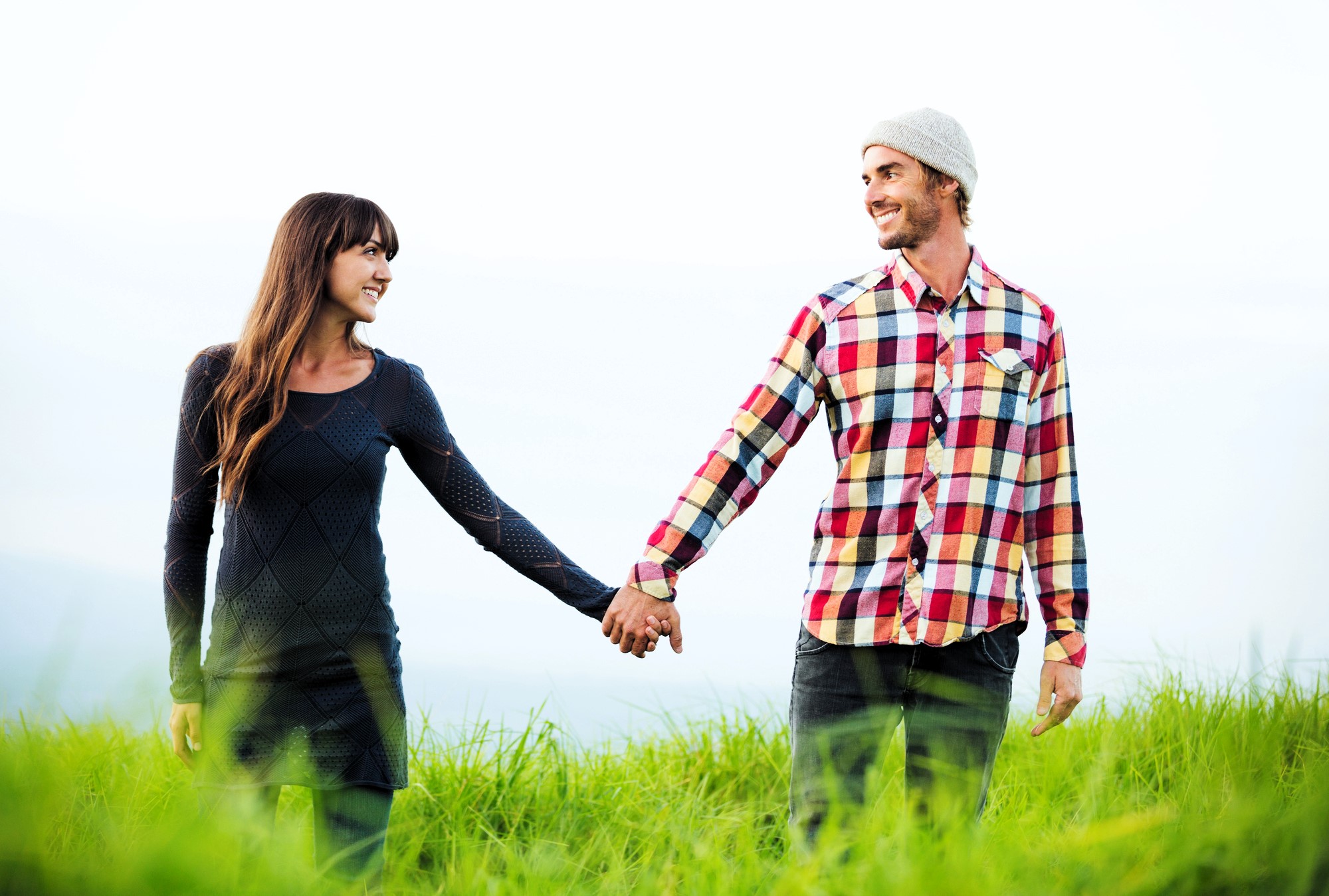 A woman in a dark dress and a man in a plaid shirt with a beanie hold hands, smiling at each other. They stand in a grassy field under a bright sky.