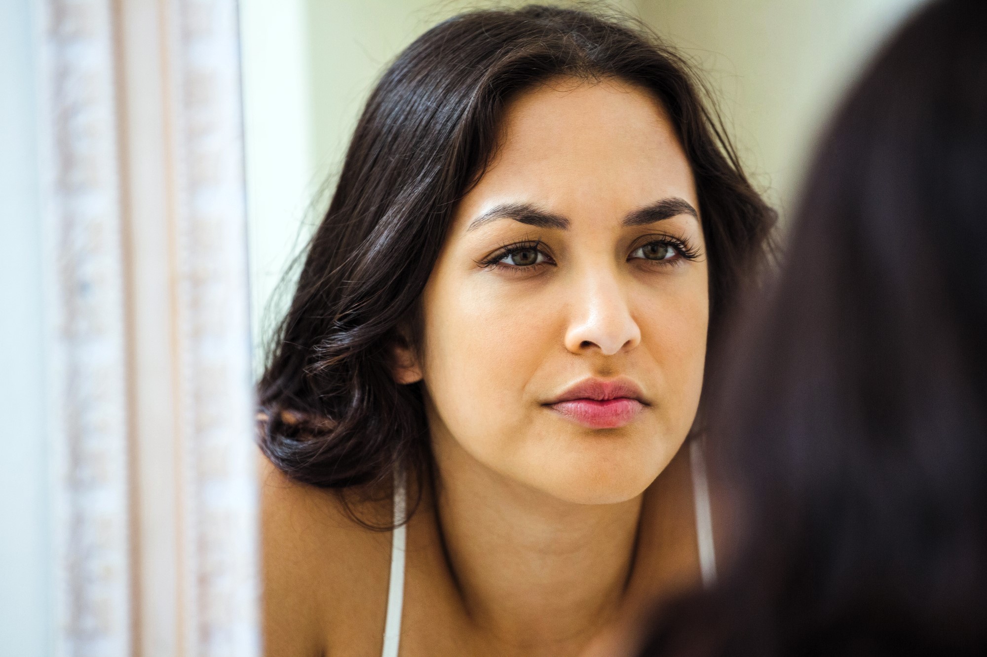 A woman with long dark hair is looking at her reflection in a mirror. She has a thoughtful expression on her face and wears a light-colored top. The setting appears to be indoors with soft lighting.