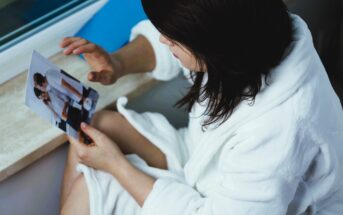 A person in a white bathrobe sits by a window, holding and looking at a photograph. The photo shows two people in a kitchen setting, one feeding the other with a spoon.