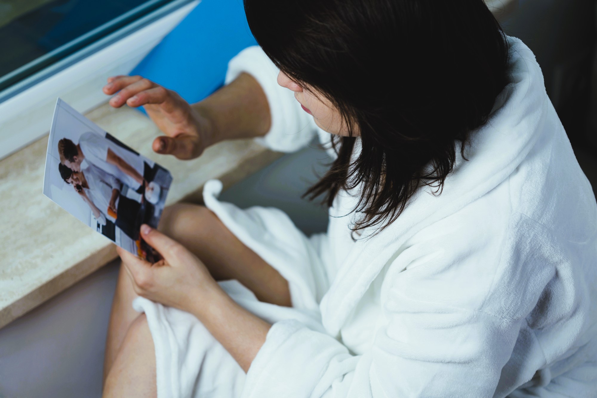 A person in a white bathrobe sits by a window, holding and looking at a photograph. The photo shows two people in a kitchen setting, one feeding the other with a spoon.