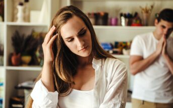 A woman with long hair, wearing a white top and shirt, looks pensive, touching her head. In the background, a man in a white shirt appears blurred, with hands pressed together. They're in a room with shelves and various items.