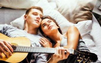 A young couple lying on a bed, surrounded by pillows. The man is playing a guitar, while the woman rests her hand on his chest, looking up. Sunlight casts a warm glow over them, creating a relaxing and intimate atmosphere.