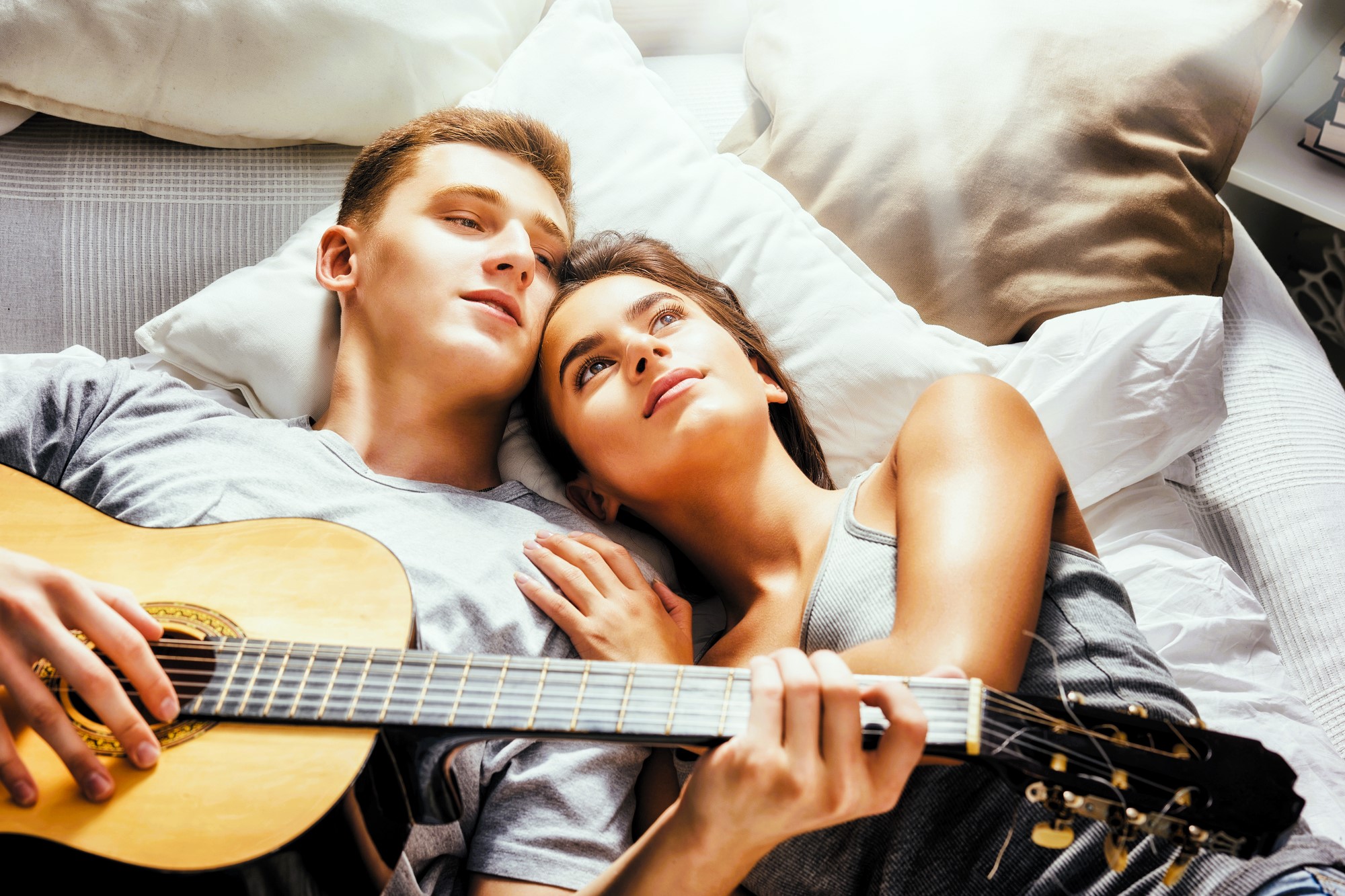 A young couple lying on a bed, surrounded by pillows. The man is playing a guitar, while the woman rests her hand on his chest, looking up. Sunlight casts a warm glow over them, creating a relaxing and intimate atmosphere.