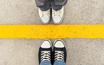 Two pairs of sneakers face each other on a concrete surface, separated by a bright yellow line. The top pair is white, and the bottom pair is black with white accents. The line creates a striking contrast against the grey background.