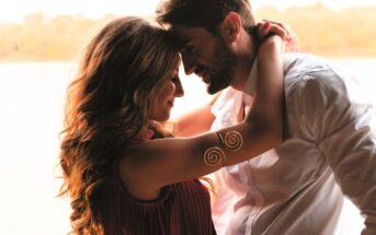 A couple facing each other closely, with the man wearing a white shirt and the woman in a sleeveless top with spiral designs on her arm. They have gentle expressions, with a blurred natural background.