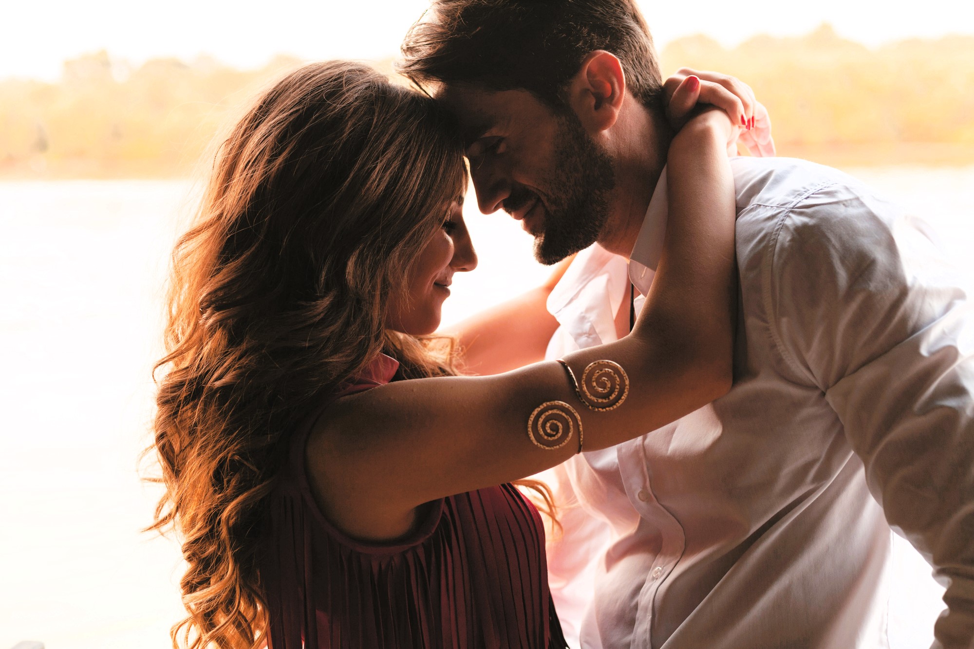 A couple facing each other closely, with the man wearing a white shirt and the woman in a sleeveless top with spiral designs on her arm. They have gentle expressions, with a blurred natural background.