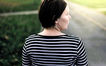 A woman with short brown hair is standing outdoors, seen from behind. She is wearing a black and white striped shirt and is facing a blurred background of a path and grass under a bright sky.