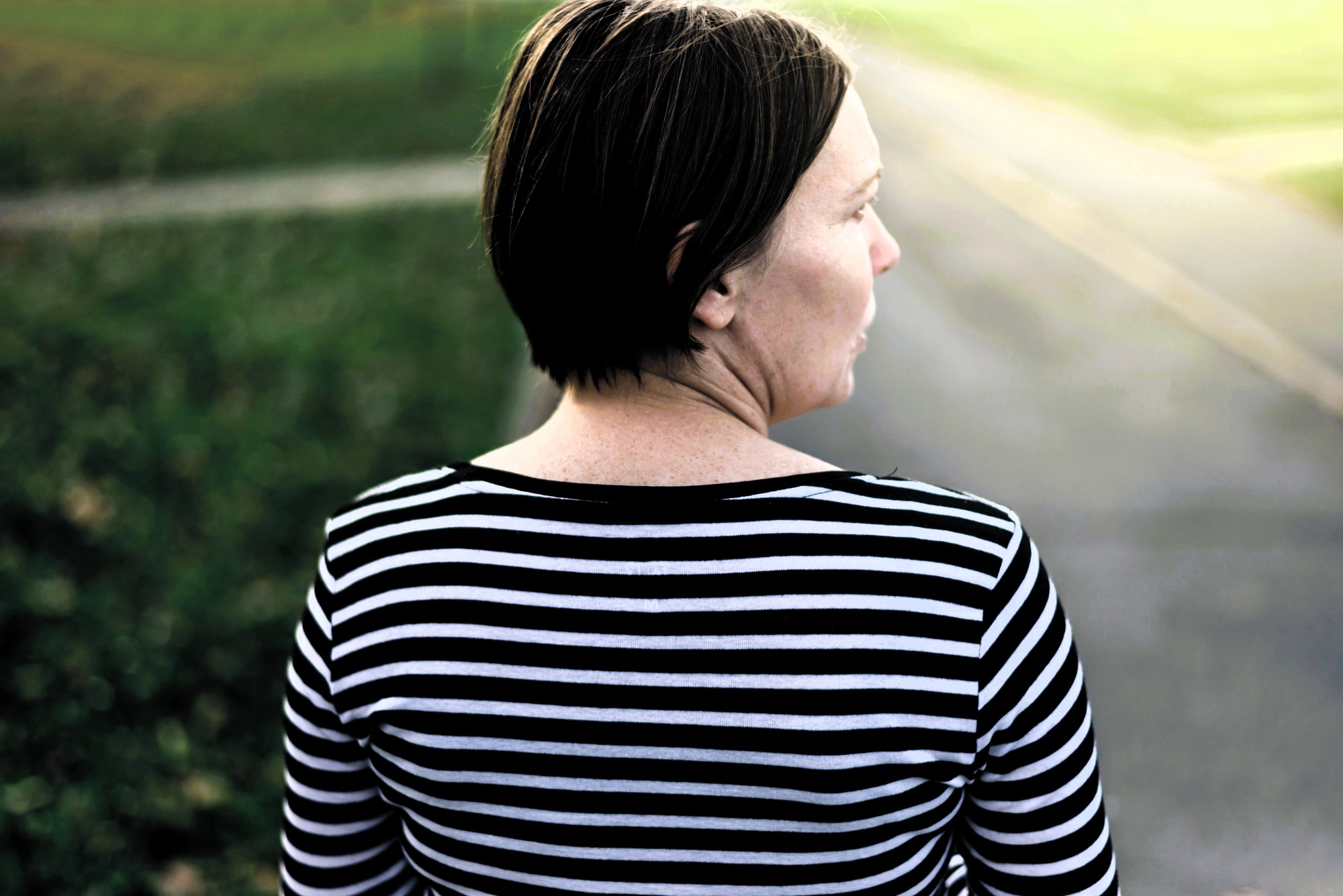 A woman with short brown hair is standing outdoors, seen from behind. She is wearing a black and white striped shirt and is facing a blurred background of a path and grass under a bright sky.