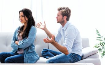 A man and woman sit on a couch. The woman has her arms crossed, looking away with a serious expression. The man gestures with his hands, appearing to speak earnestly. Both are wearing casual shirts and jeans. A plant is visible beside them.