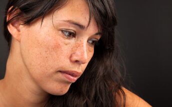 A woman with long brown hair and freckles looks down, slightly away from the camera, against a dark background. She appears thoughtful or contemplative.