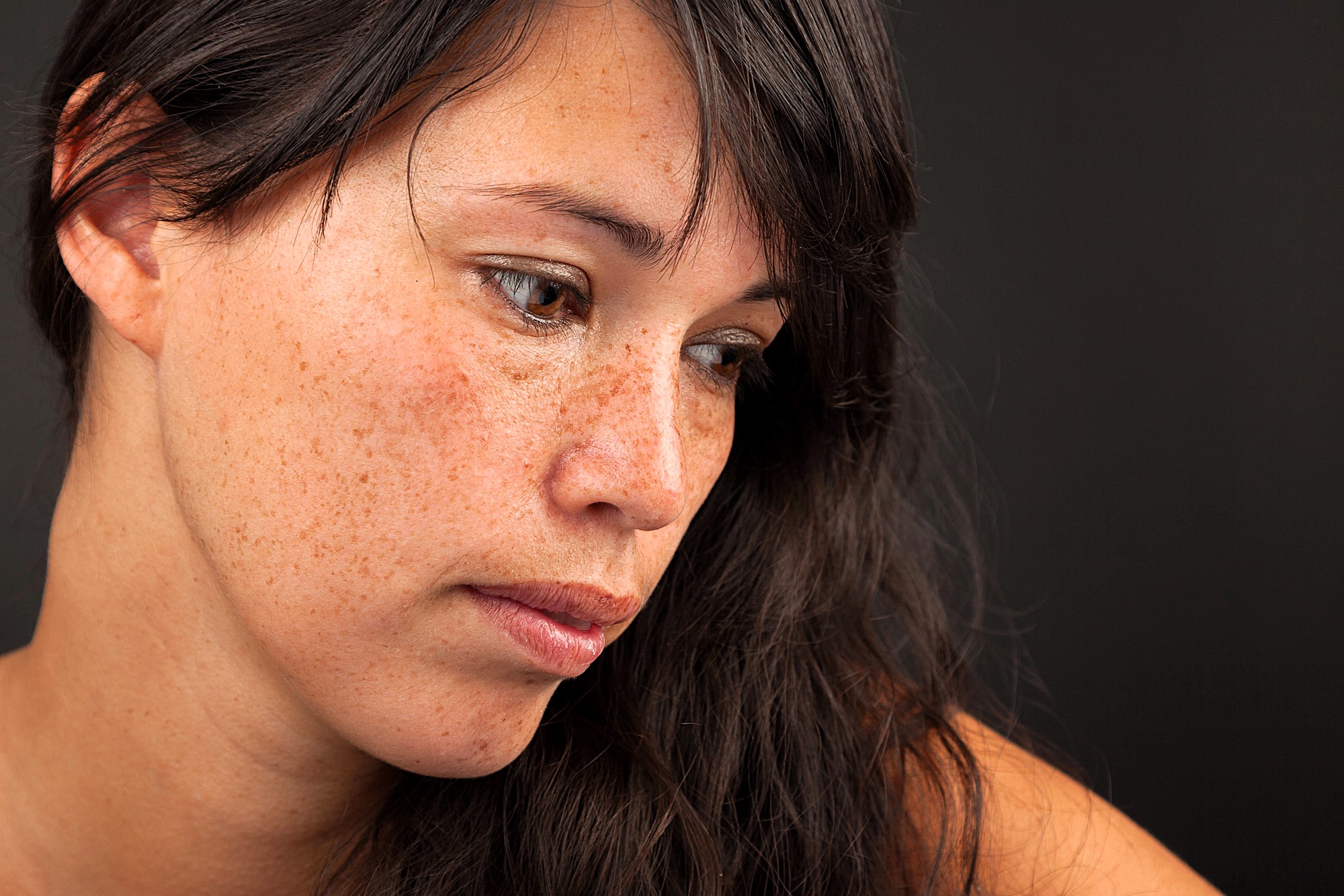 A woman with long brown hair and freckles looks down, slightly away from the camera, against a dark background. She appears thoughtful or contemplative.