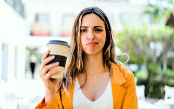 A person with long hair, wearing a yellow jacket and white top, holds a coffee cup while making a puzzled expression. The background is blurred with greenery and buildings.