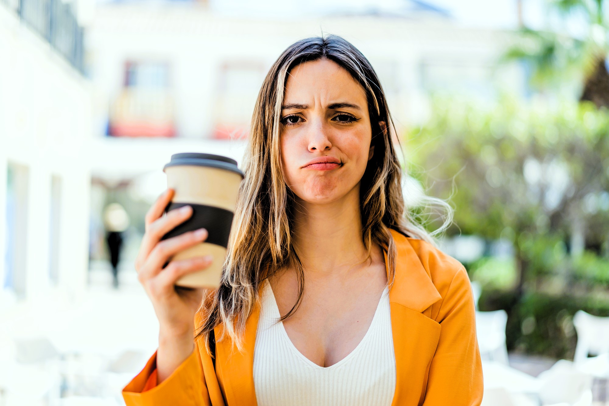 A person with long hair, wearing a yellow jacket and white top, holds a coffee cup while making a puzzled expression. The background is blurred with greenery and buildings.