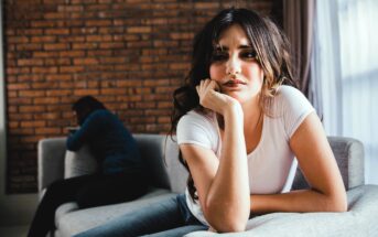 A woman with long hair, wearing a white t-shirt, is leaning forward on a gray sofa, looking thoughtful. In the background, another person is sitting with their head down on a cushion, facing away. The room features a brick wall.