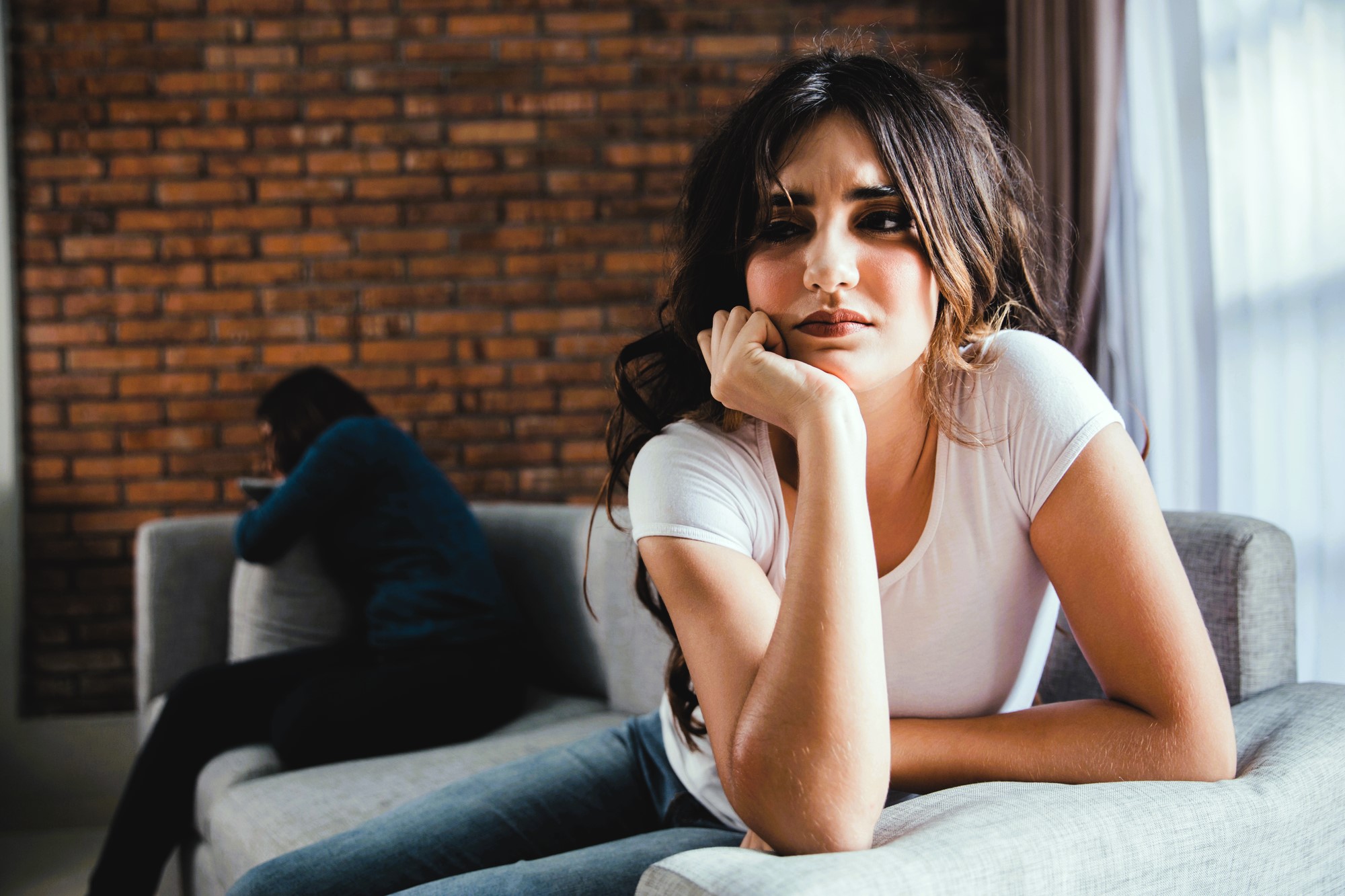 A woman with long hair, wearing a white t-shirt, is leaning forward on a gray sofa, looking thoughtful. In the background, another person is sitting with their head down on a cushion, facing away. The room features a brick wall.