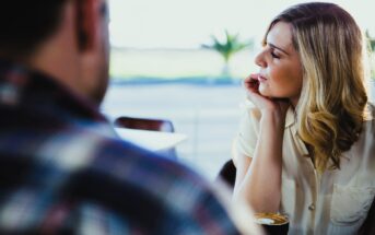 A woman with blonde hair, wearing a white shirt, sits at a table in a café with a cup of coffee. She rests her chin on her hand and gazes thoughtfully out the window. Another person, blurred, is in the foreground wearing a plaid shirt.