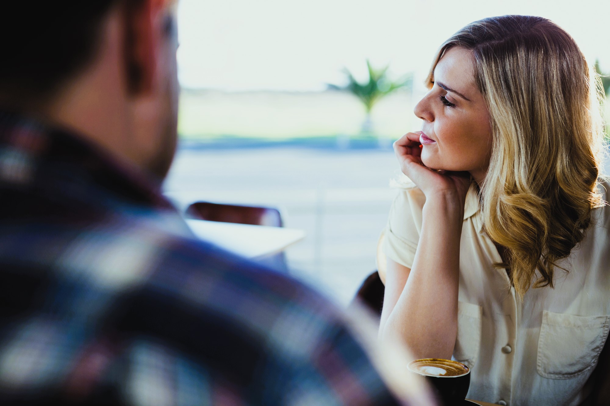 A woman with blonde hair, wearing a white shirt, sits at a table in a café with a cup of coffee. She rests her chin on her hand and gazes thoughtfully out the window. Another person, blurred, is in the foreground wearing a plaid shirt.