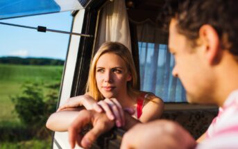 A woman and a man sit together in a van parked in a scenic area. The woman is leaning on the window, looking thoughtfully outside, while the man looks towards her. Outside, there is greenery and an open field under a clear blue sky.