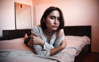 A person with short brown hair is lying on a bed, looking at the camera. They are wearing a lace top and a knitted cardigan, with a choker necklace. The room has a mirror, a dresser, and warm lighting.