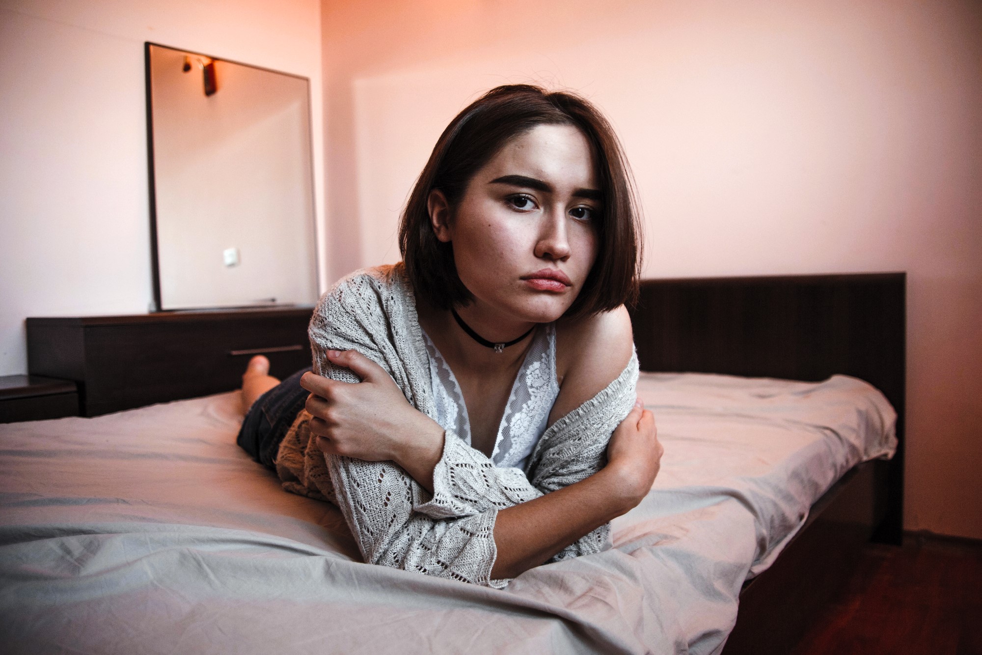 A person with short brown hair is lying on a bed, looking at the camera. They are wearing a lace top and a knitted cardigan, with a choker necklace. The room has a mirror, a dresser, and warm lighting.