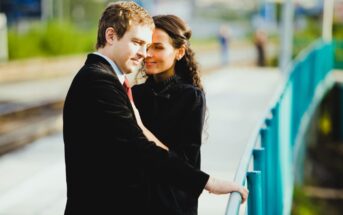 A couple stands close together on a walkway, sharing an intimate moment. The man is dressed in a suit and red tie, while the woman wears a black outfit. They lean against a teal railing, with blurred greenery and structures in the background.