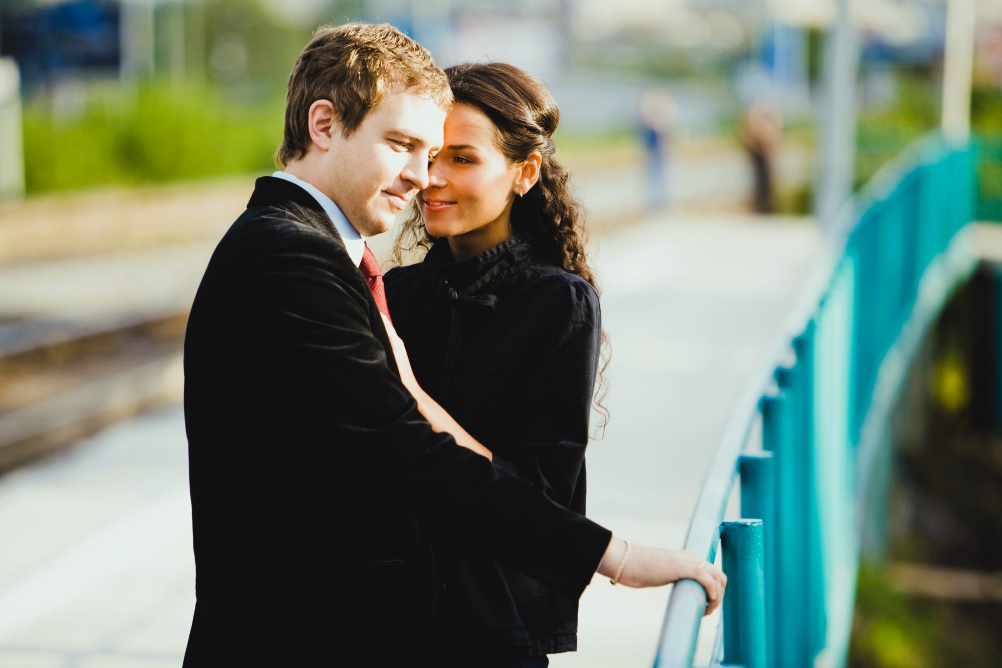 A couple stands close together on a walkway, sharing an intimate moment. The man is dressed in a suit and red tie, while the woman wears a black outfit. They lean against a teal railing, with blurred greenery and structures in the background.