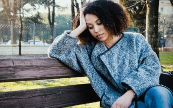 A woman with curly hair wearing a gray coat sits on a wooden bench in a park, resting her head on one hand. She appears thoughtful or pensive. Trees and a fence are visible in the background.