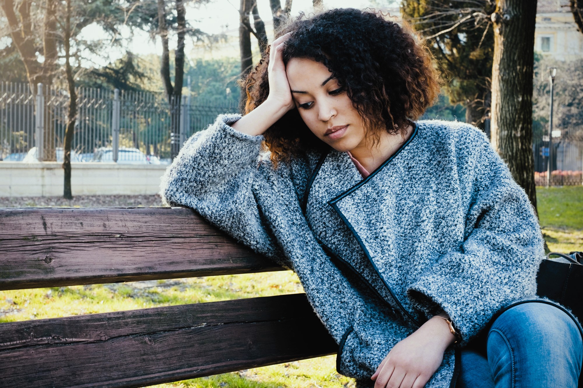 A woman with curly hair wearing a gray coat sits on a wooden bench in a park, resting her head on one hand. She appears thoughtful or pensive. Trees and a fence are visible in the background.