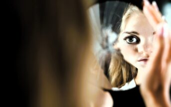 A woman with long blonde hair is looking into a cracked mirror, her face partially visible. One eye is prominently reflected in a shard. The background is dark, highlighting the fragmented reflection.