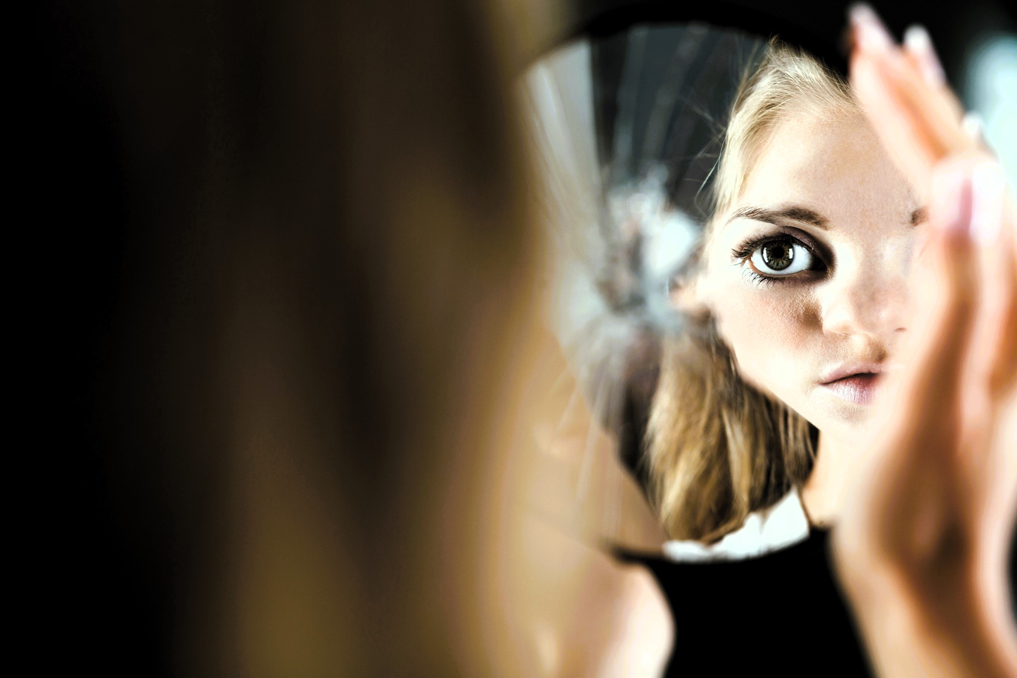 A woman with long blonde hair is looking into a cracked mirror, her face partially visible. One eye is prominently reflected in a shard. The background is dark, highlighting the fragmented reflection.