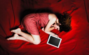 A woman with curly hair lies on a red blanket, wearing a red dress. She is curled up on her side with her head resting on one arm and is holding a tablet with a blank screen in her other hand.