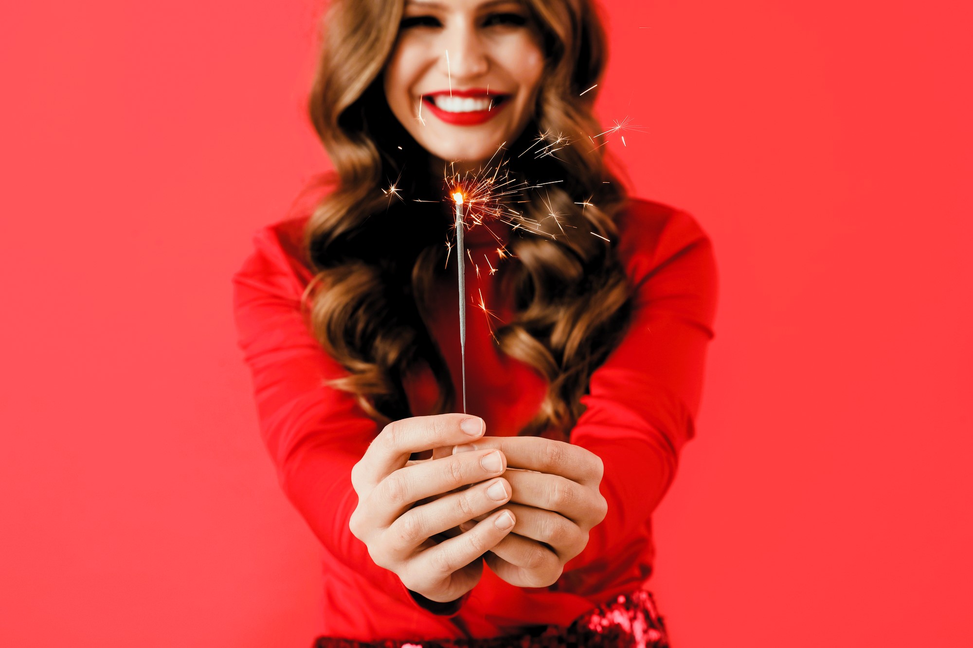 A woman with long, wavy brown hair smiles widely while holding a lit sparkler in both hands. She is wearing a red outfit, and the background is a matching vibrant red color.