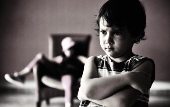 A young boy with an unhappy expression stands cross-armed in the foreground, wearing a striped shirt. In the blurred background, there is a person sitting in a chair with their legs crossed, wearing a cap.