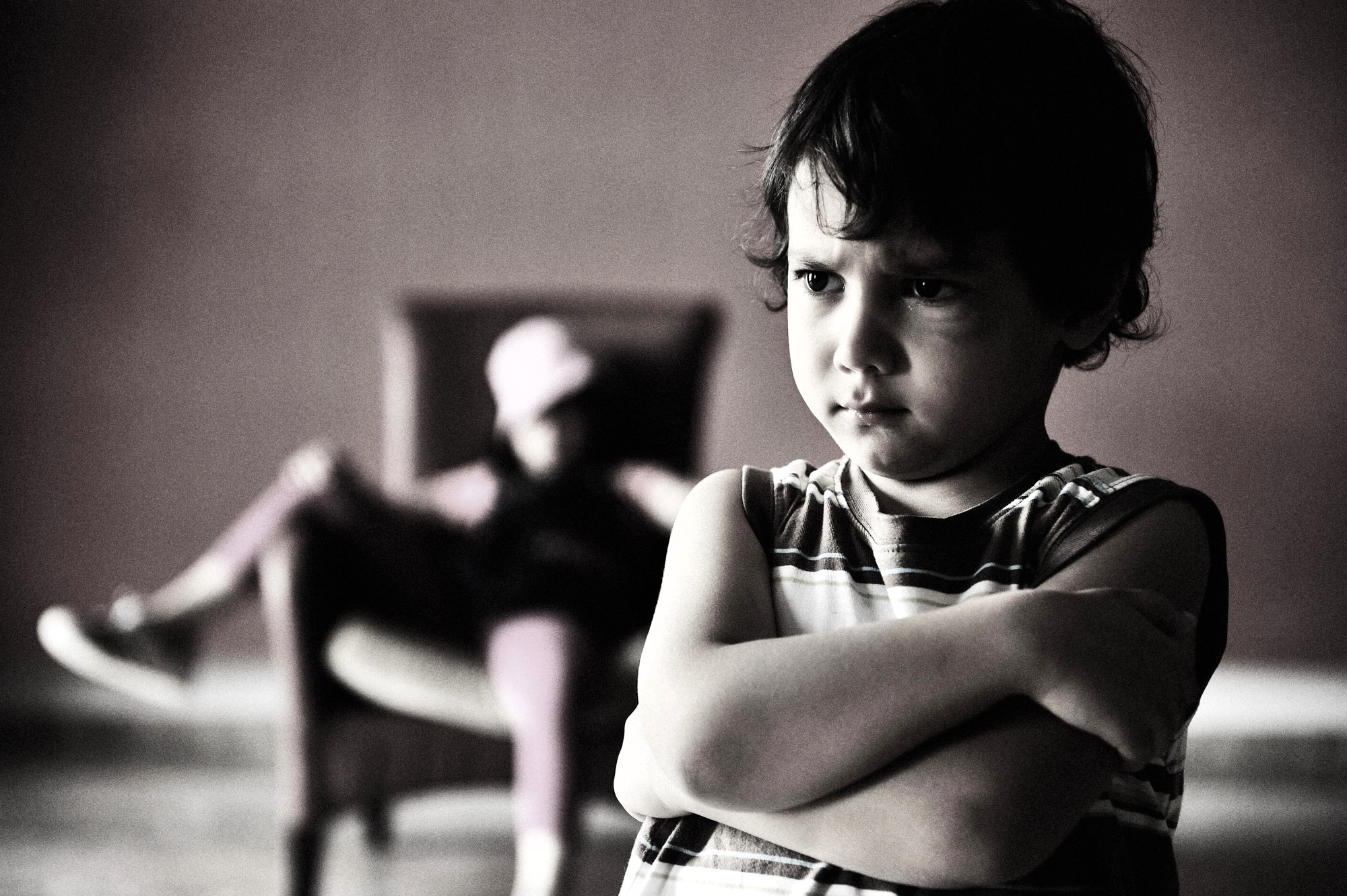 A young boy with an unhappy expression stands cross-armed in the foreground, wearing a striped shirt. In the blurred background, there is a person sitting in a chair with their legs crossed, wearing a cap.